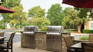 Een terras buiten met twee roestvrijstalen barbecues, stenen muren en rieten stoelen.