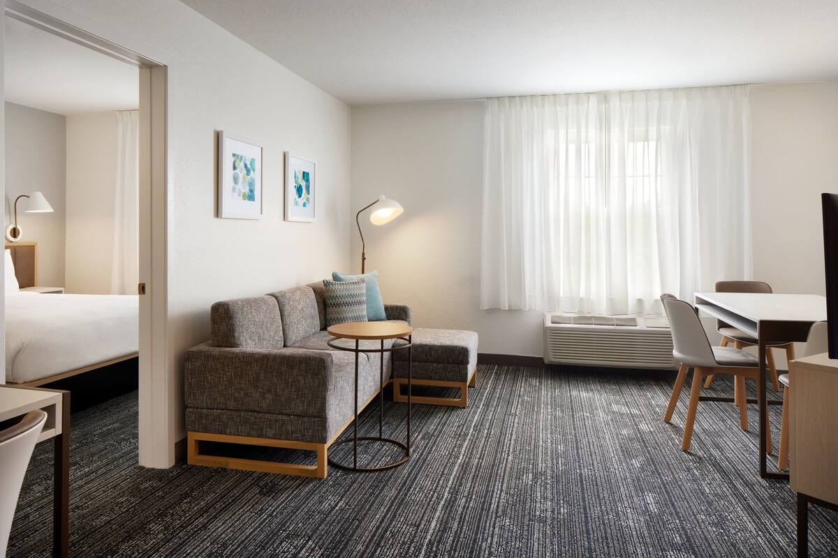 Hotellihuoneessa on sänky, harmaa sohva, pieni pyöreä pöytä, työpöytä ja tuoleja. Seinällä on kaksi kehystettyä kuvaa.