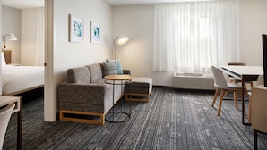 A hotel room with a bed, a gray sofa, a small round table, a desk, and chairs. There are two framed pictures on the wall.