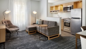 A modern living room with a sofa, armchair, and round table. A kitchen with a microwave, oven, and refrigerator is visible in the background.
