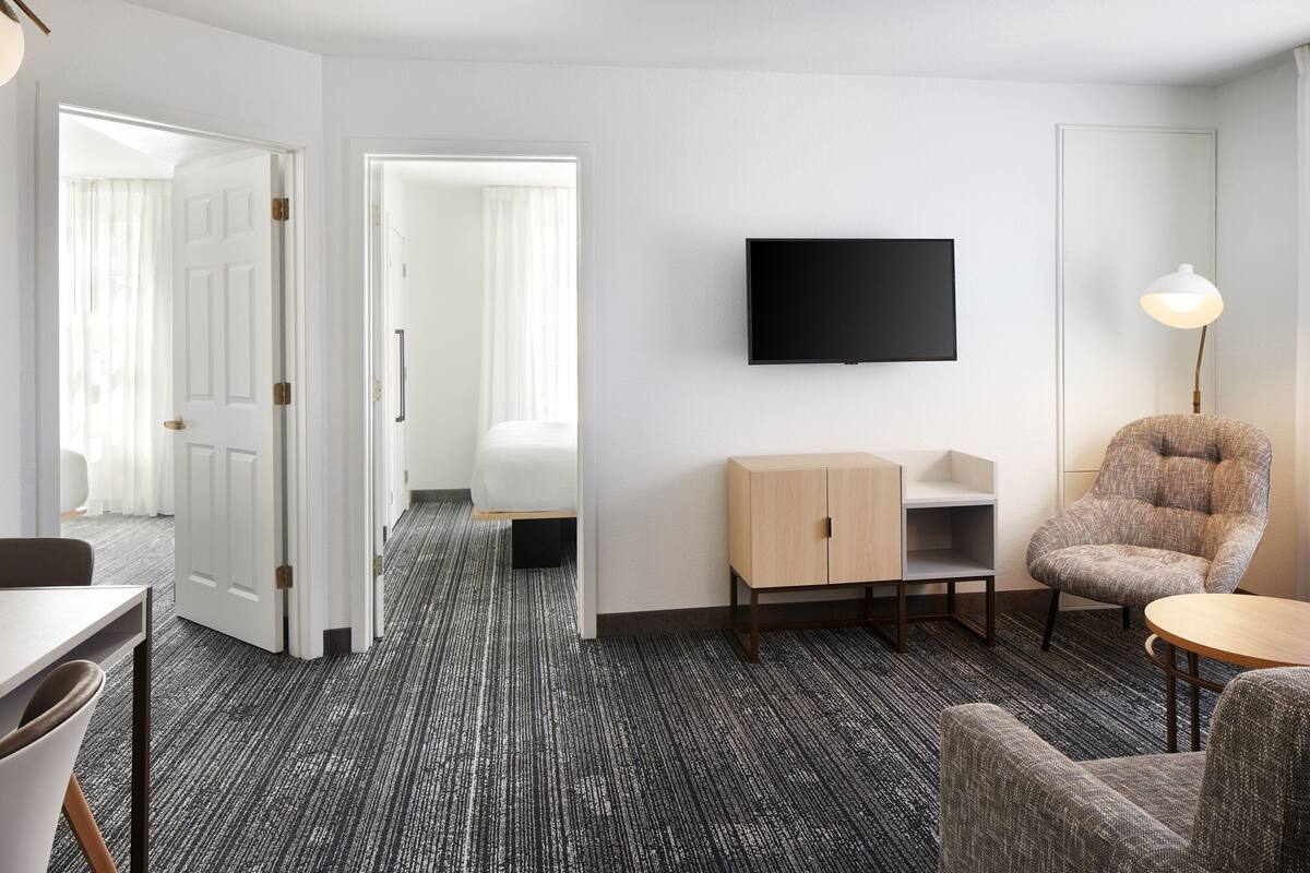 Hotellihuone, jossa on seinälle asennettu taulutelevisio, puinen kaappi, harmaa nojatuoli ja pieni pyöreä pöytä.