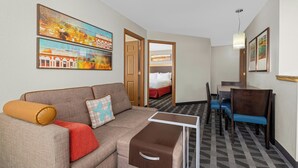 Habitación de hotel con sofá, una pequeña mesa, zona de comedor con sillas, cama y obras de arte en las paredes.