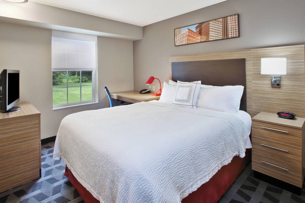A hotel room with a large bed, a desk with a chair, a television, a window with a view of greenery, and a painting on the wall.