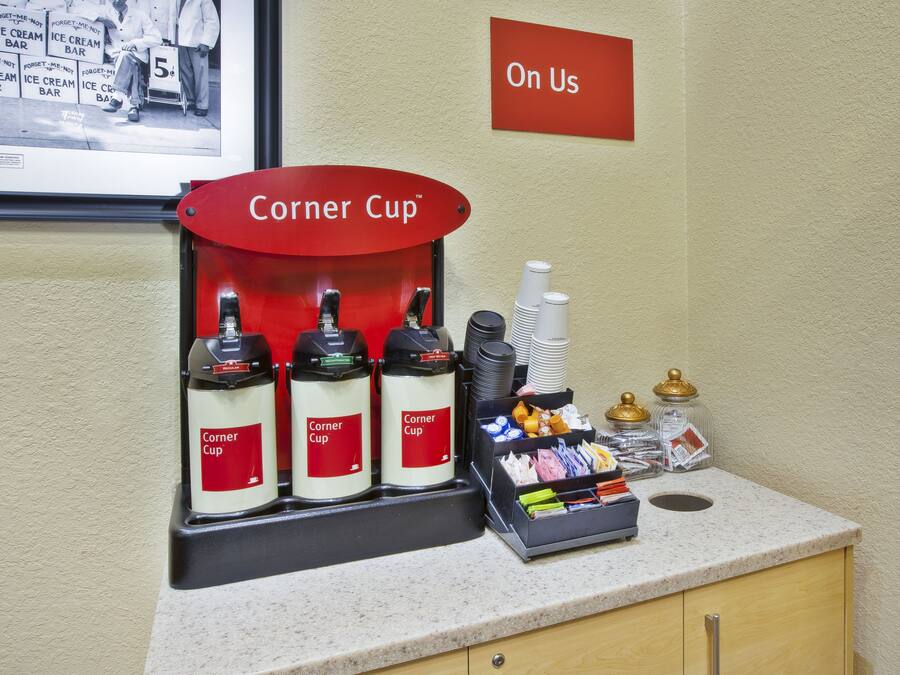 Una estación de bebidas con tres dispensadores Corner Cup, una pila de vasos desechables y una bandeja con condimentos y servilletas.