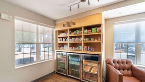 A corner of a room with a wooden shelf unit stocked with snacks and beverages, a leather armchair, and a glass-fronted mini-fridge. - Extended Stay America Suites - Norfolk - Virginia Beach (Virginia Beach)