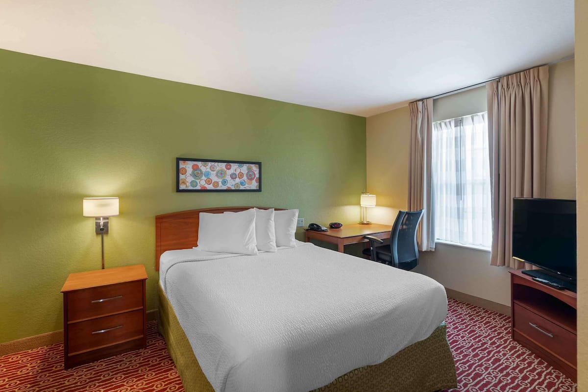 A hotel room with a bed, bedside tables, a desk, a chair, a television, and a window with curtains.