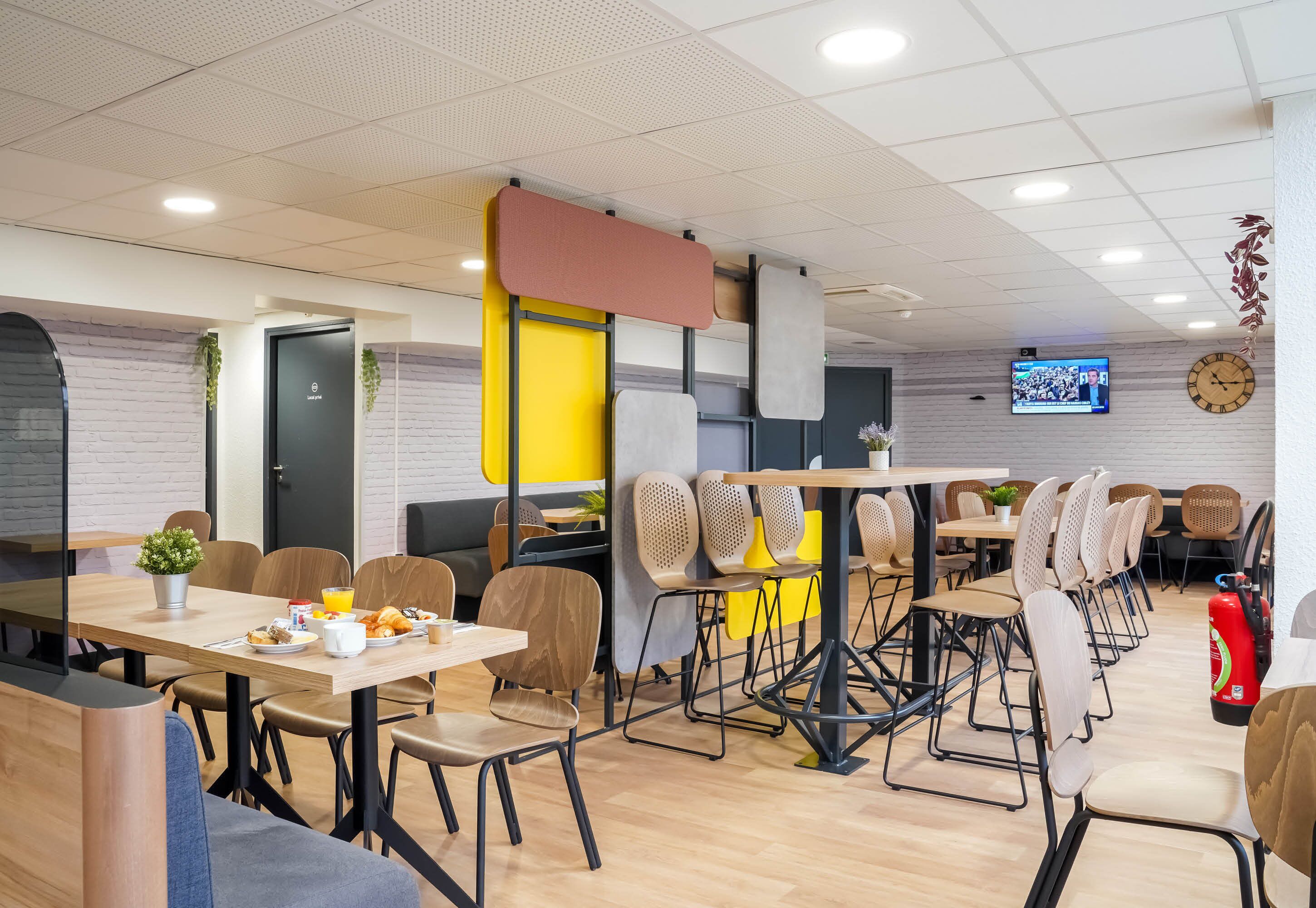 A modern dining area with multiple tables and chairs, a clock on the wall, and a television screen.