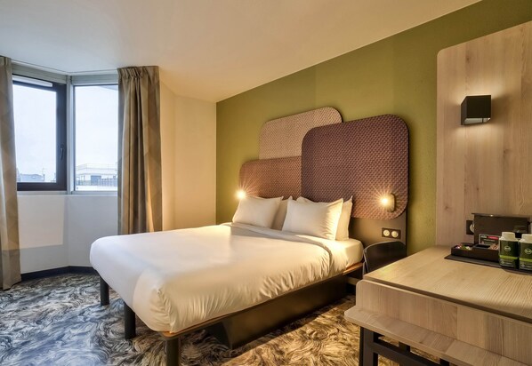A hotel room with a large bed, a desk with a coffee machine, and a view of the city through the window.