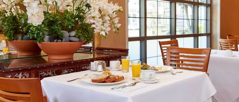 Una escena de desayuno con una mesa preparada para dos personas, incluyendo platos de comida, un vaso de jugo de naranja y un jarrón con flores blancas.