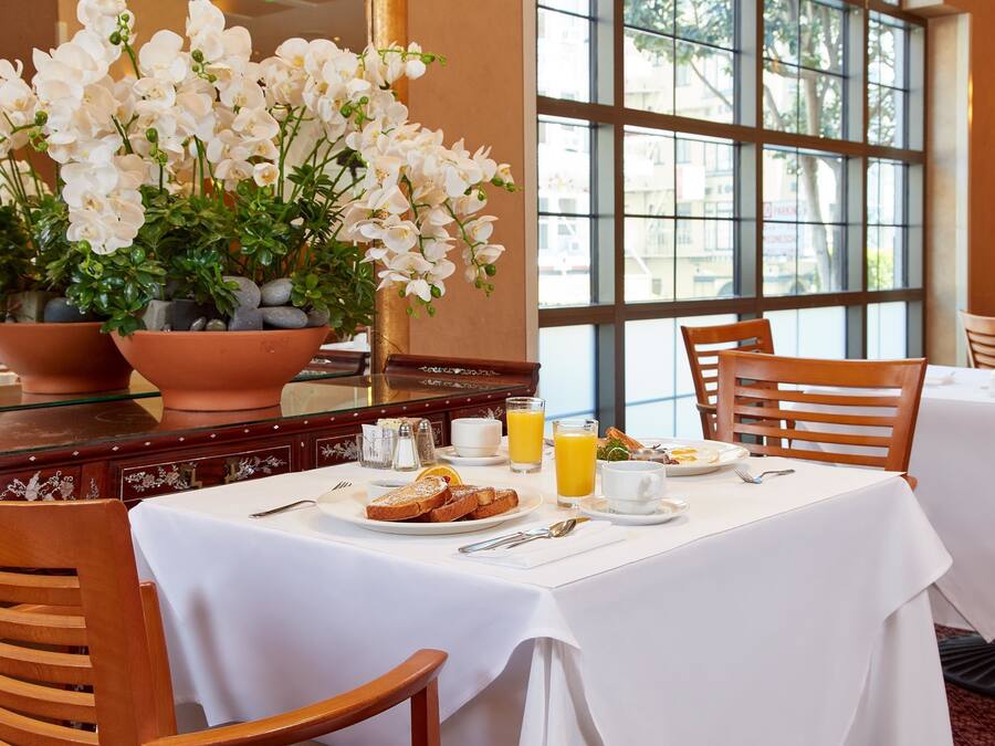 A breakfast setting with a table set for two, including plates of food, a glass of orange juice, and a vase of white flowers.
