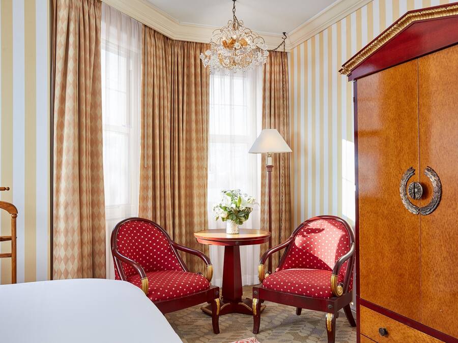 Hotelkamer met een bed, twee rode fauteuils, een houten kast, een ronde tafel met een lamp en een kroonluchter.