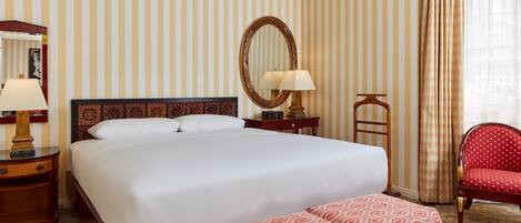 A hotel room with a large bed, a bedside table with a lamp, a mirror, a chair, and a patterned bench.
