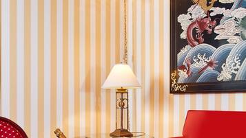 A room with striped wallpaper, a red sofa, a chandelier, a framed artwork, a table with a bottle of champagne, a lamp, and a tray of chocolate-covered strawberries.