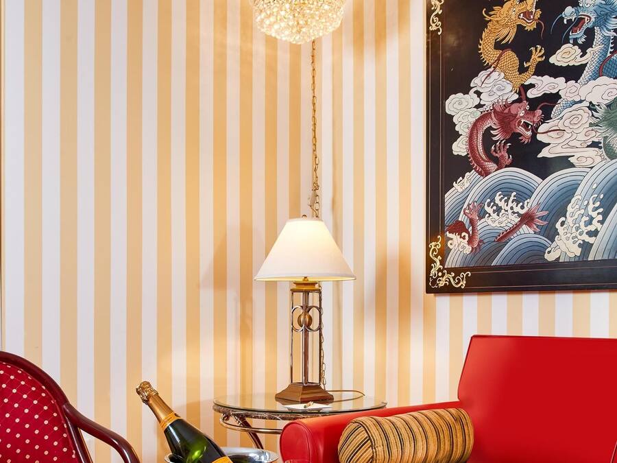 Een kamer met gestreepte behang, een rode bank, een kroonluchter, een ingelijste kunstwerk, een tafel met een fles champagne, een lamp en een schaal met chocoladebedekte aardbeien.