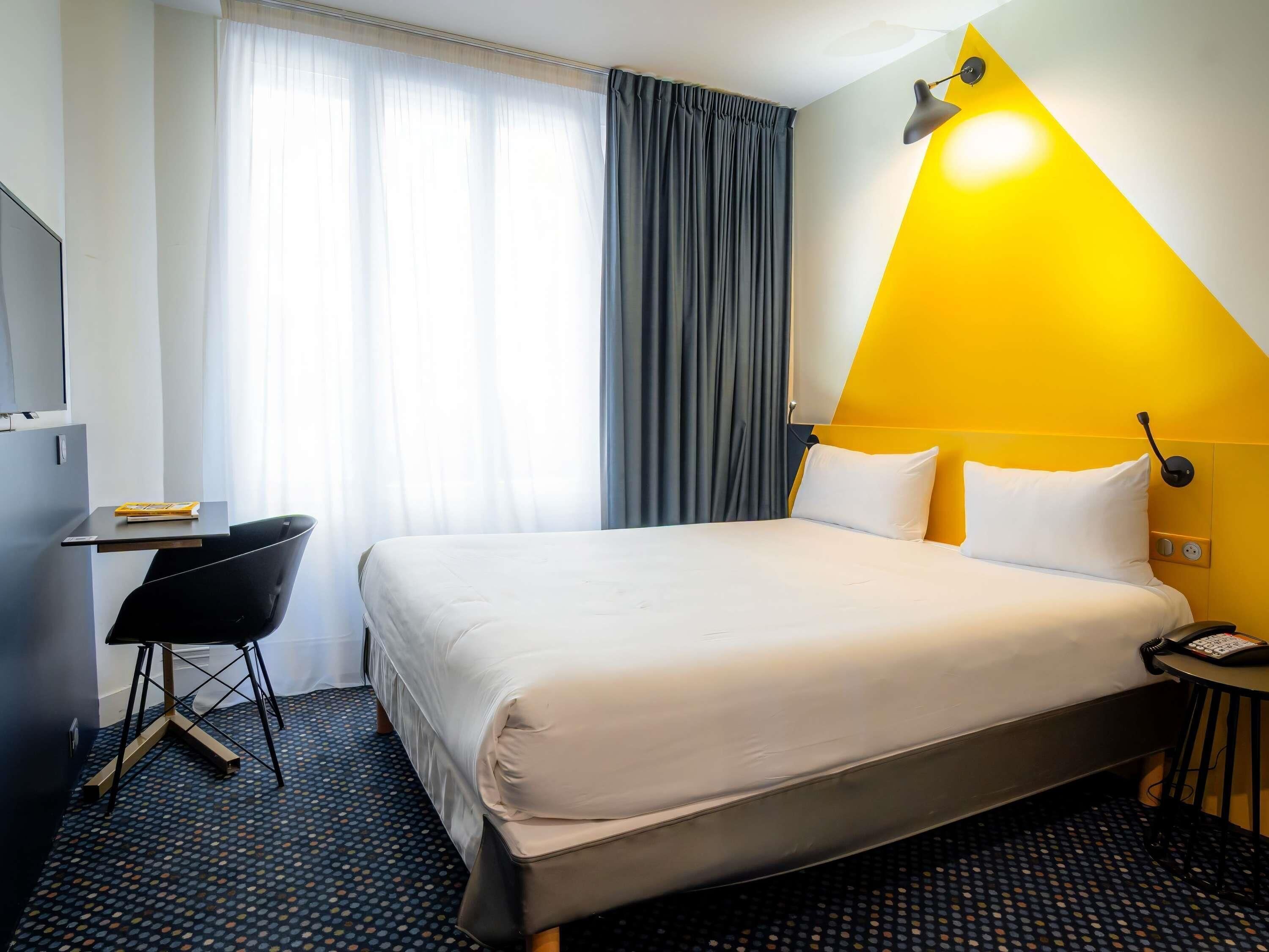 A hotel room with a bed, a desk, a chair, a lamp, and a phone.