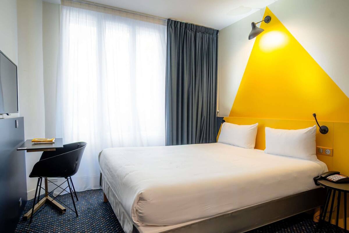 A hotel room with a bed, a desk, a chair, a lamp, and a phone.