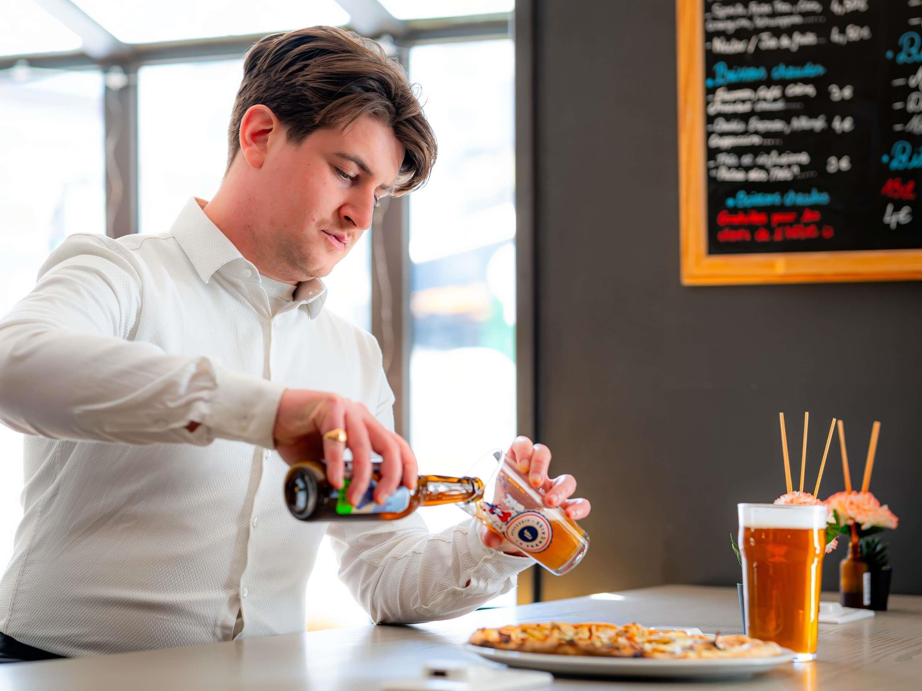 A person pouring beer into a glass, a pizza on the table, and a menu in the background.