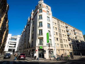 Um edifício de vários andares com uma placa verde grande, entrada com portão e carros estacionados.