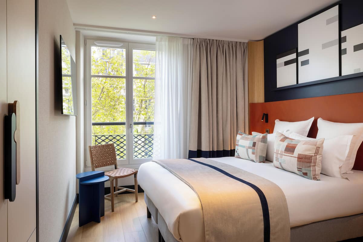 A hotel room with a large bed, a chair, a small table, and a window with curtains.