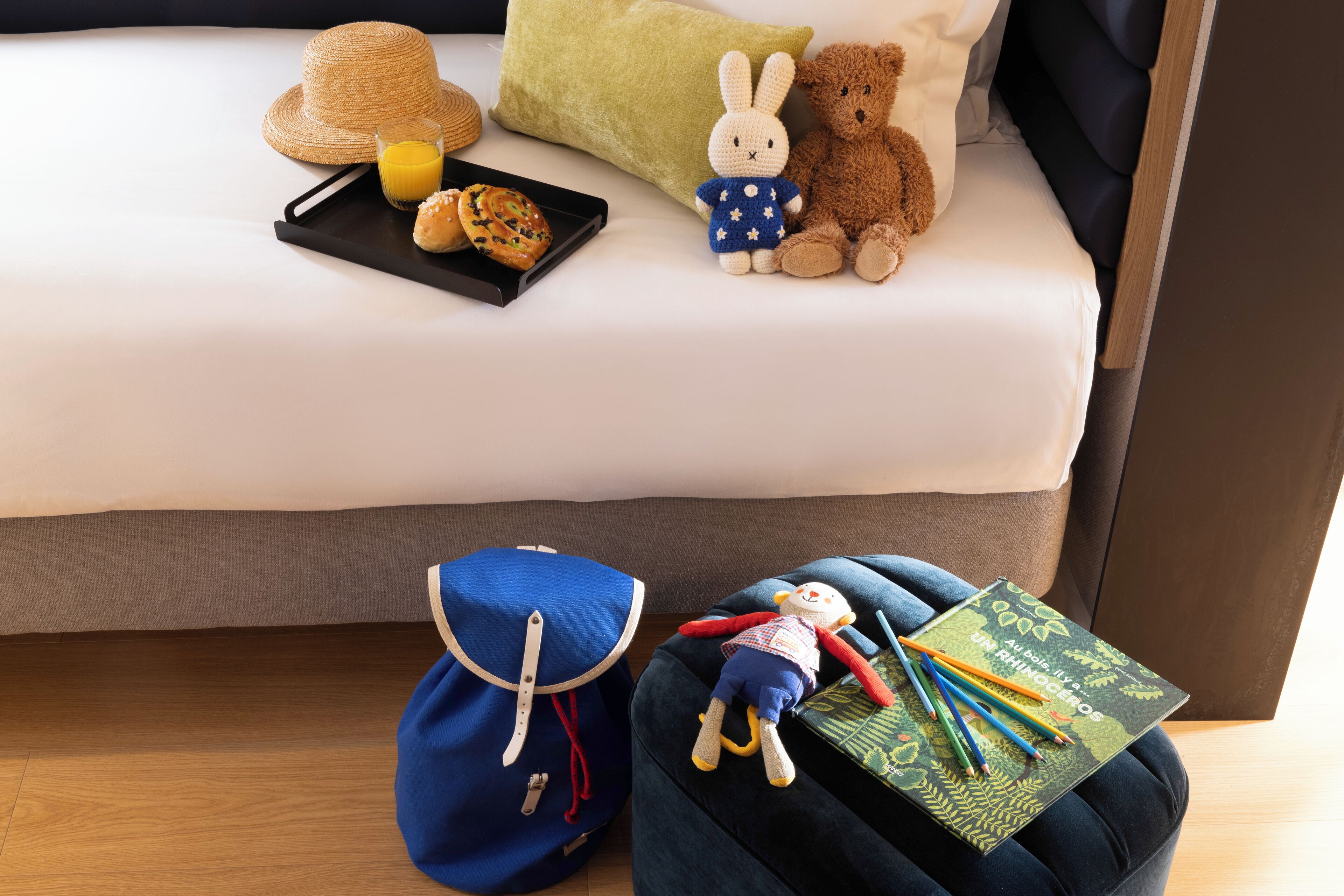Una cama ordenada con un sombrero de paja, un peluche, un osito de peluche y una bandeja con bollería y zumo.
