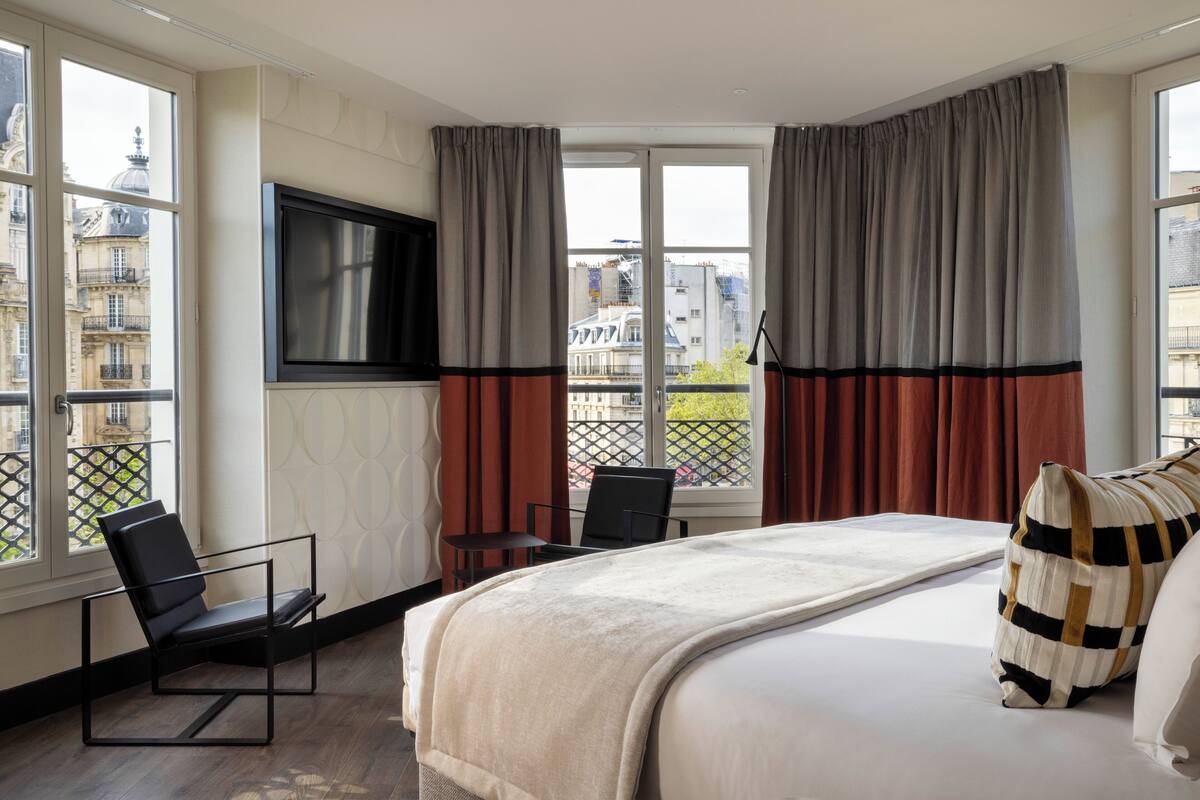 A hotel room with a large bed, a TV mounted on the wall, and a view of the city through the windows.