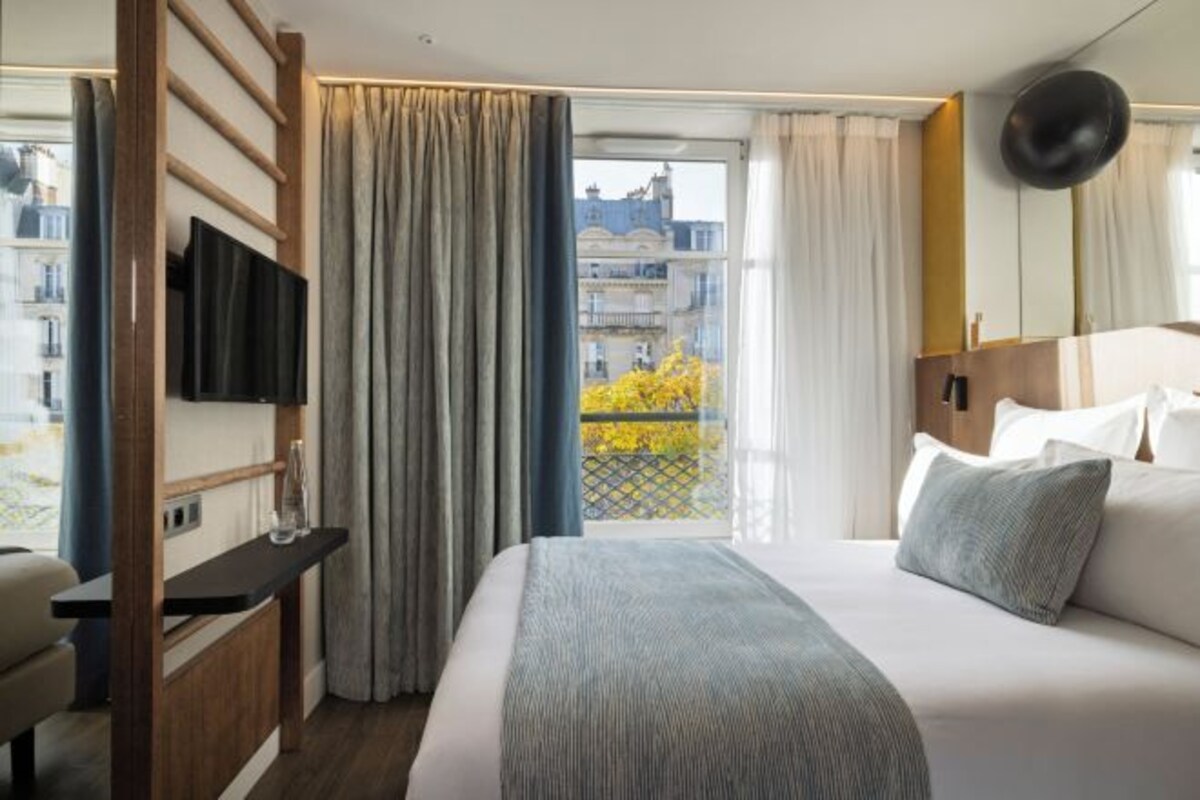 Una habitación de hotel con cama, escritorio, televisión y balcón con cortinas.