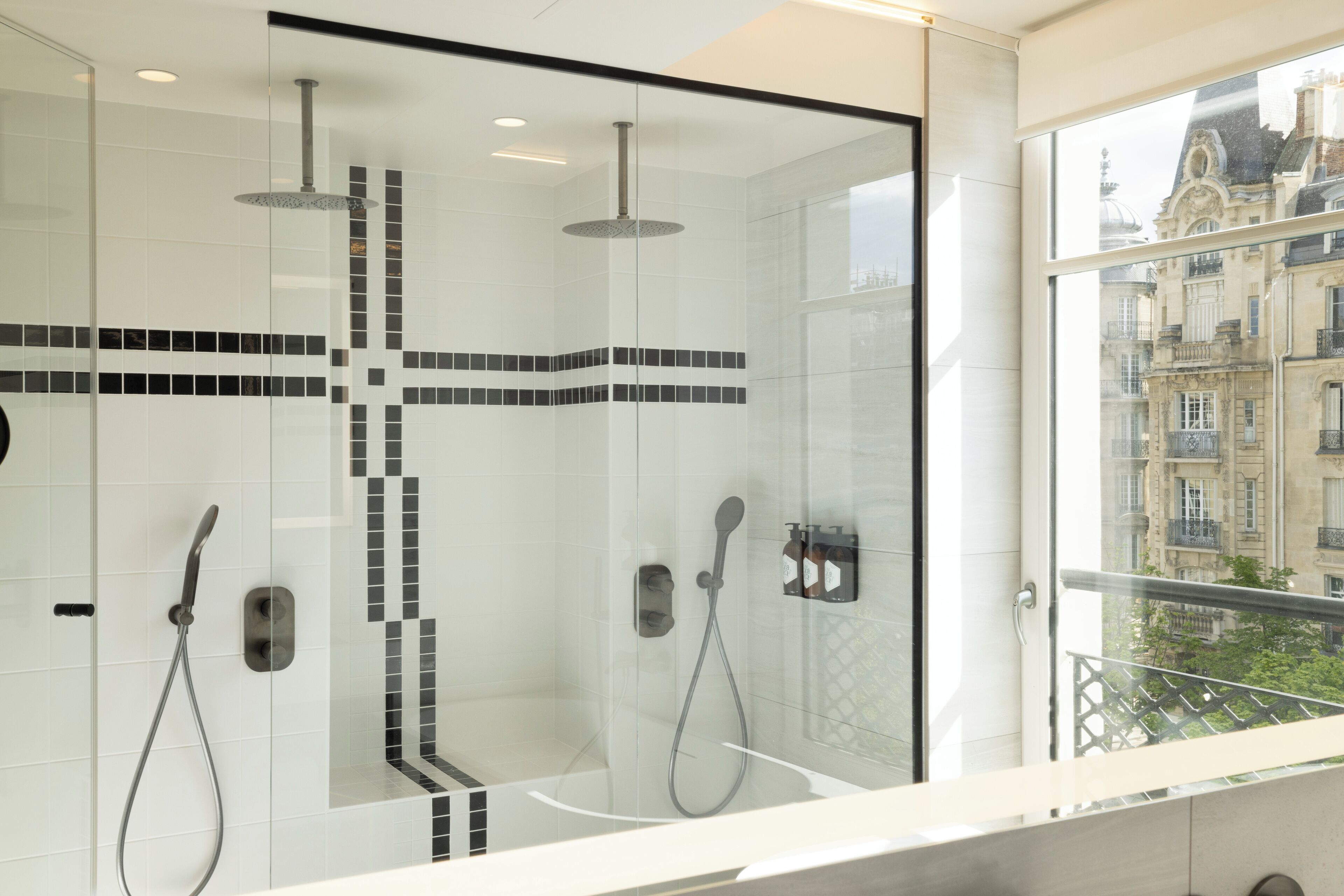 Een moderne badkamer met een glazen douchecabine, een witte betegelde muur met zwarte accenten, en uitzicht op historische gebouwen door een groot raam.