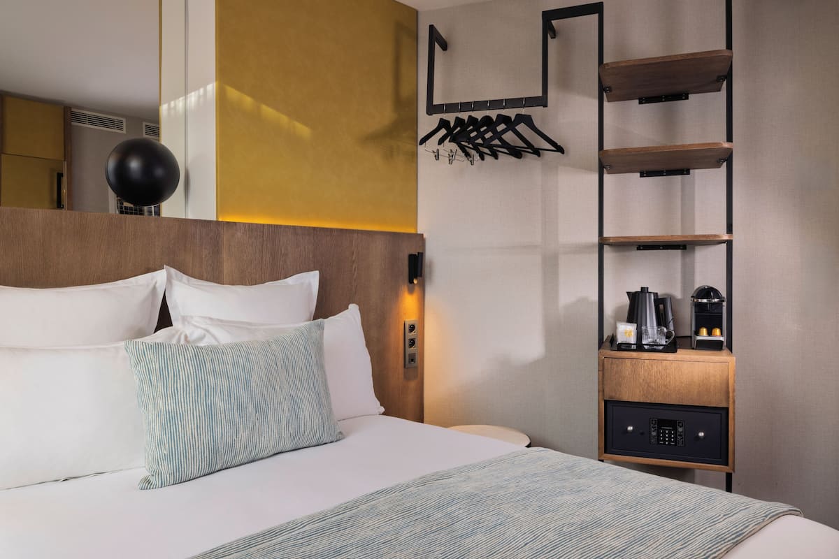 A hotel room with a bed, a bedside table with a coffee maker, a wall-mounted shelf, and a hanging coat rack.
