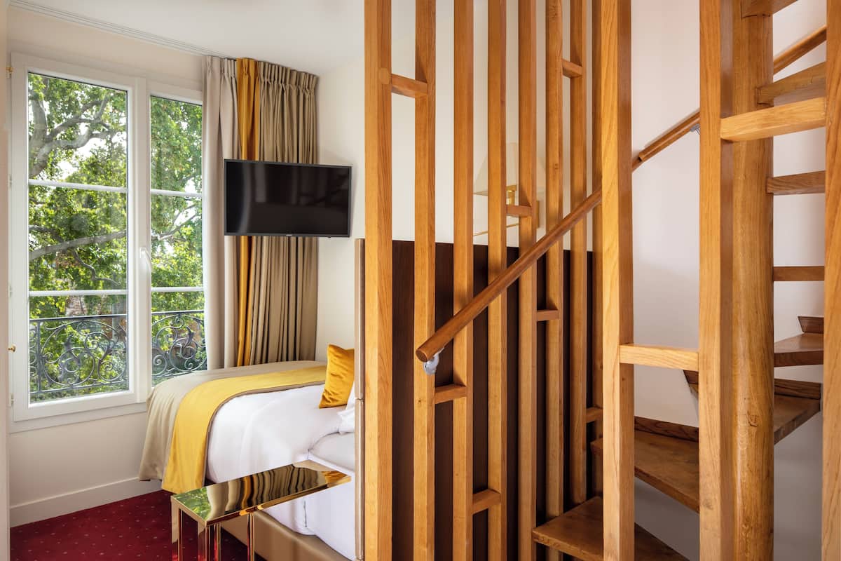 A bedroom with a bed, a TV mounted on the wall, a window with curtains, and a wooden staircase.