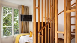A bedroom with a bed, a TV mounted on the wall, a window with curtains, and a wooden staircase.