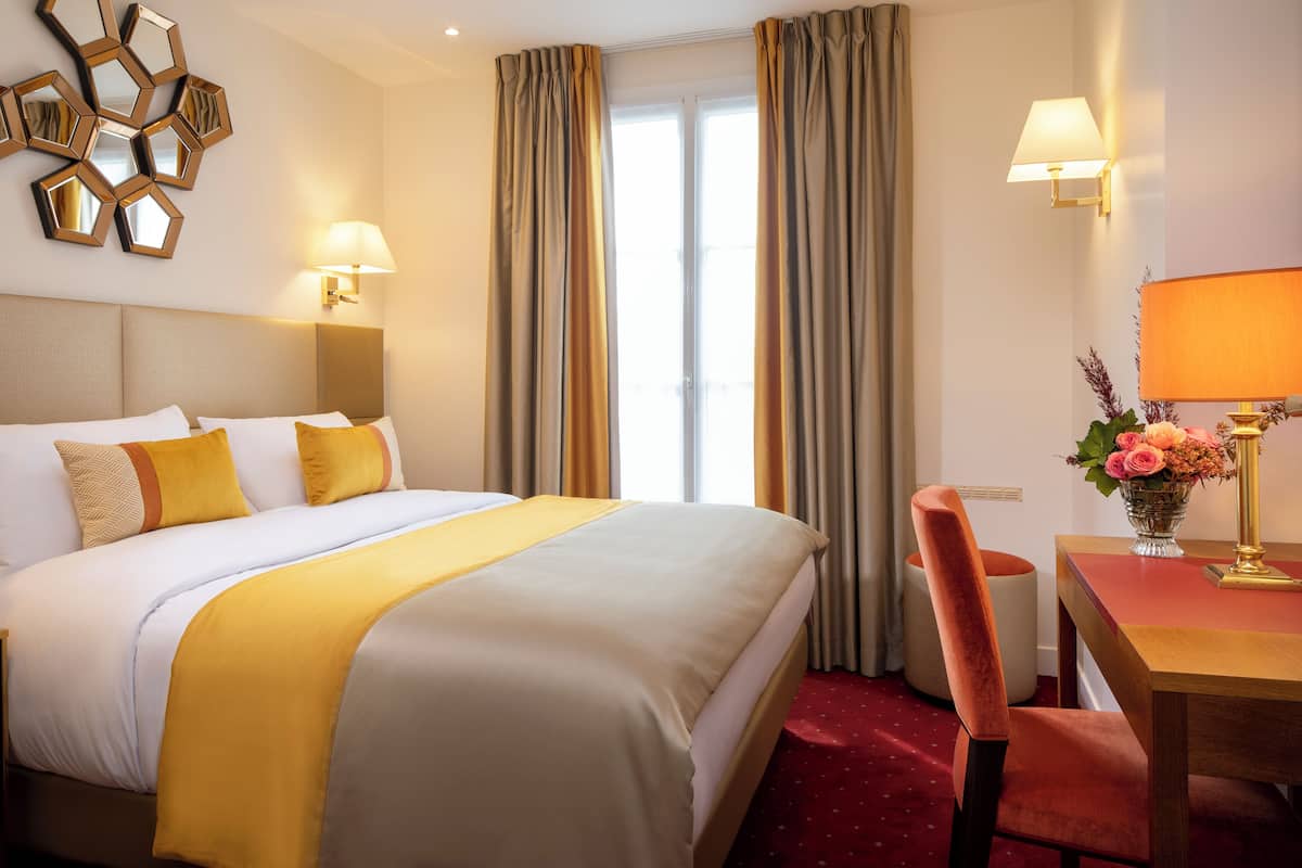 A hotel room with a large bed, two bedside lamps, a desk with a chair, and a window with curtains.