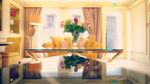 A room with a glass table reflecting the interior, a yellow sofa, a vase with flowers, and a lamp.