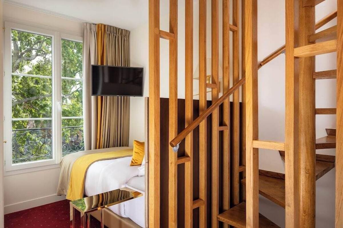 A room with a bed, a TV mounted on the wall, a window with curtains, and a wooden staircase.