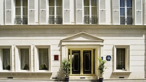 A hotel facade with a symmetrical design, featuring a central entrance with double doors, flanked by windows with shutters, and potted plants at the entrance.