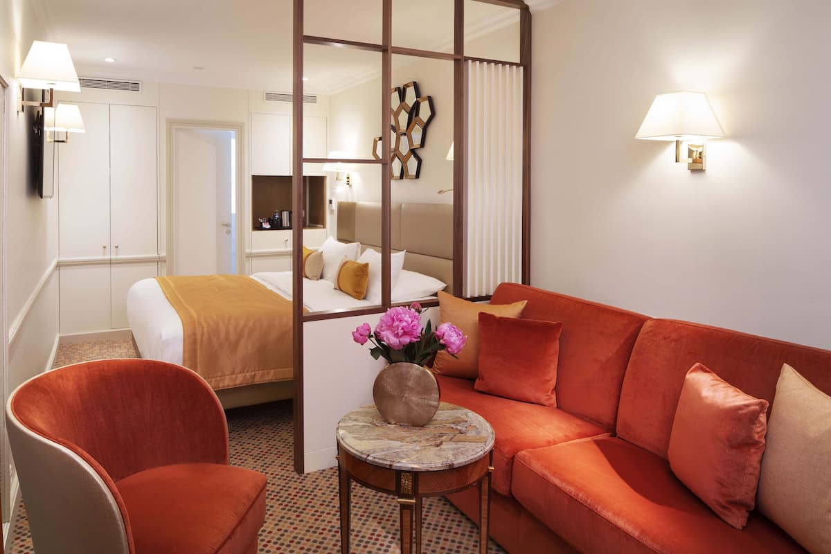 A hotel room with a bed, a sofa, a chair, a small table with a vase of flowers, and wall-mounted lamps.