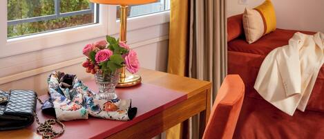 Habitación de hotel con un escritorio de madera, una lámpara, un florero con flores, una cama con ropa de cama roja y una ventana con cortinas.