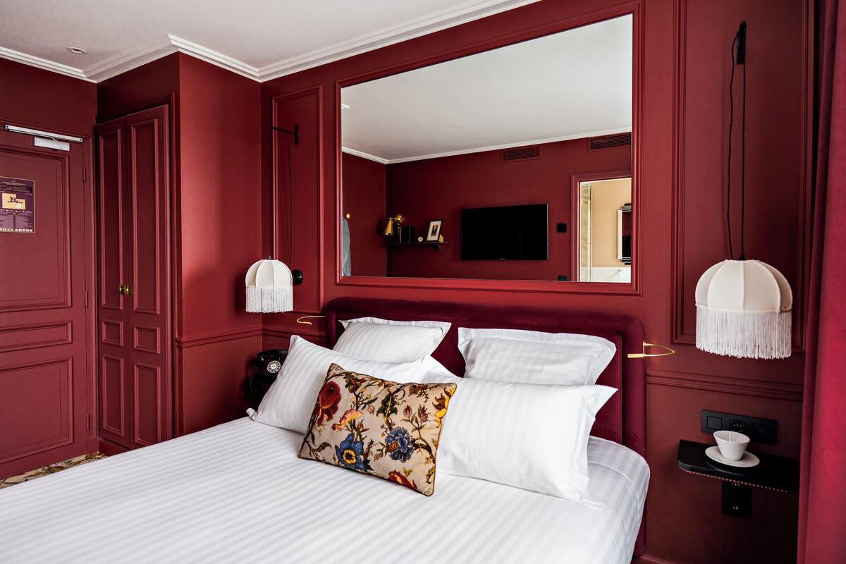 A room with a large bed, a mirror, a television, and a bedside table with a lamp.