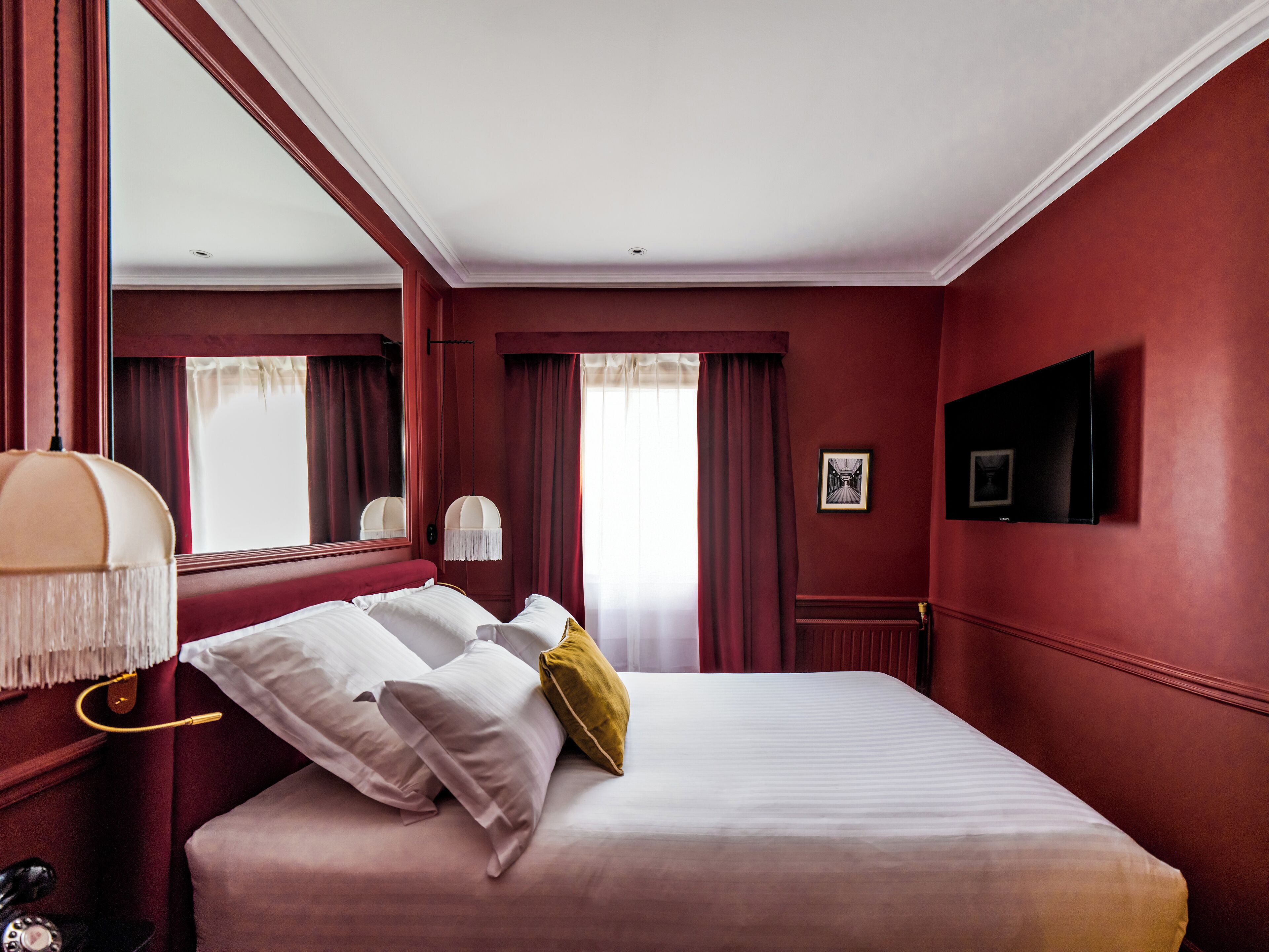 Una habitación de hotel con una cama grande, una mesita de noche con lámpara, un televisor montado en la pared y una ventana con cortinas.