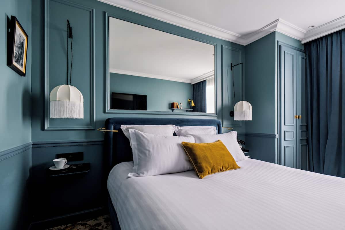 A neatly arranged hotel room with a large bed, a bedside table with a cup and saucer, a mirror above the bed, and a dark blue wall.