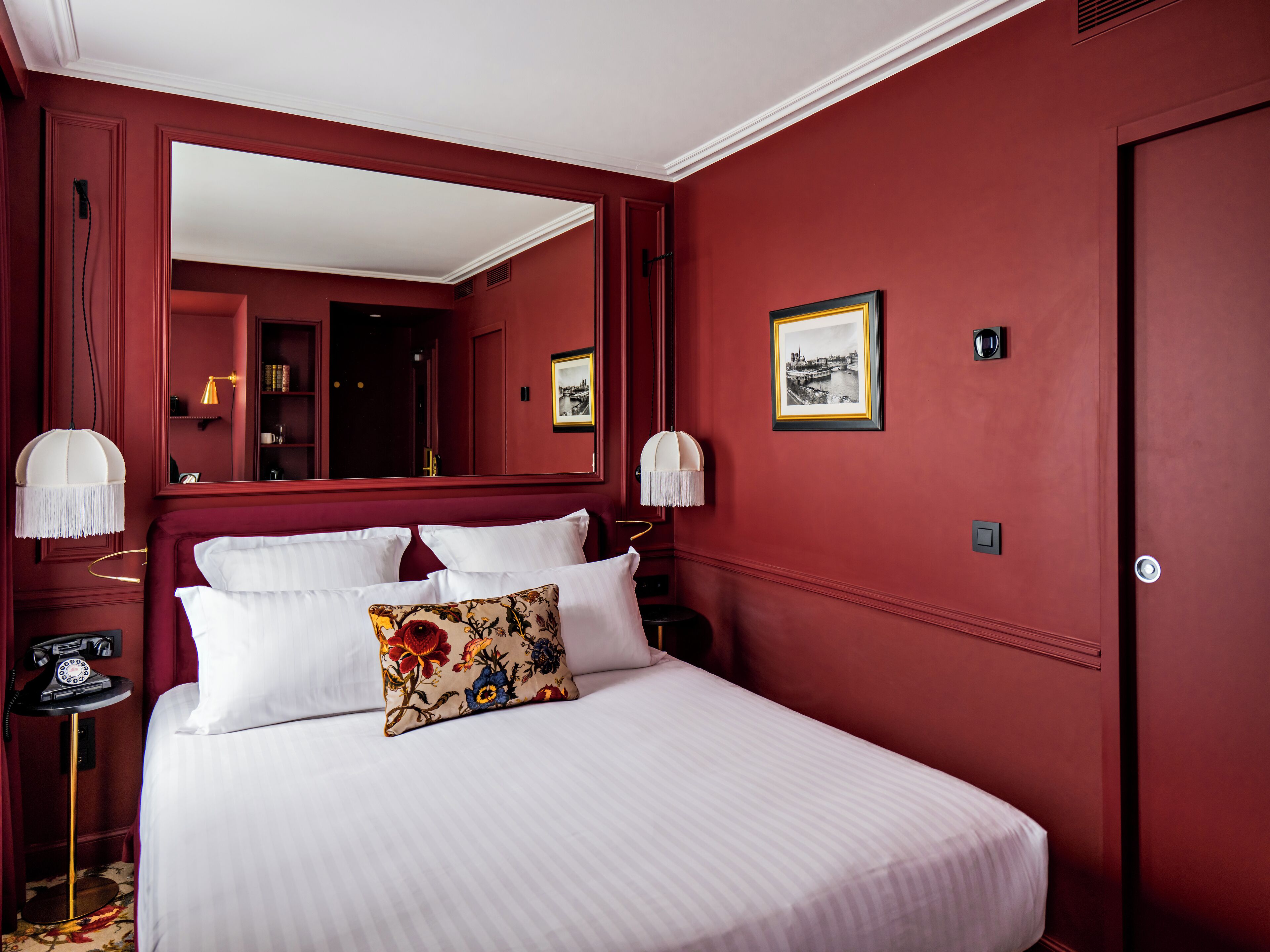 A hotel room with a large bed, white bedding, a red accent wall, a mirror above the bed, and a framed picture on the wall.