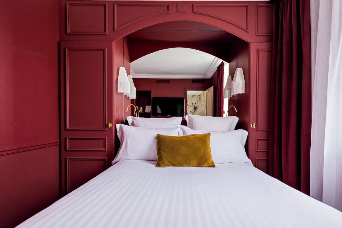 A room with a large bed, white linens, a mustard-colored pillow, and a red wall.