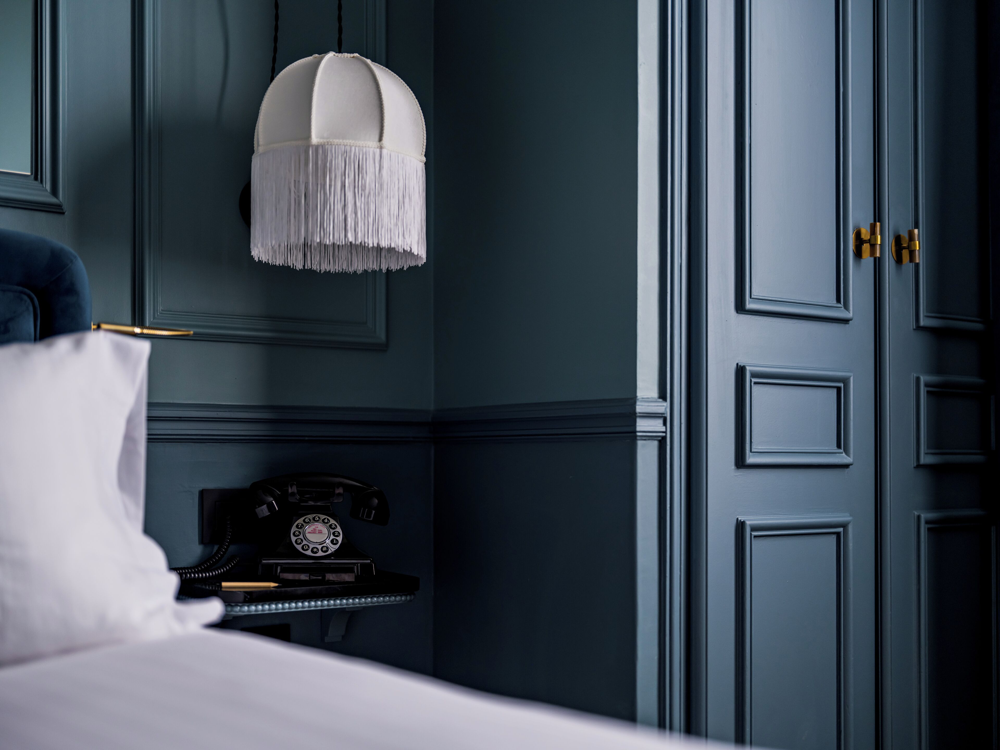 A room with a bed, a nightstand with a rotary phone, a white lamp, and a blue door.