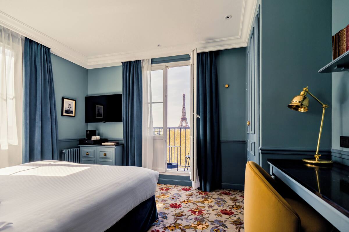 A hotel room with a bed, a desk, a chair, a desk lamp, a dresser, and a view of the Eiffel Tower.
