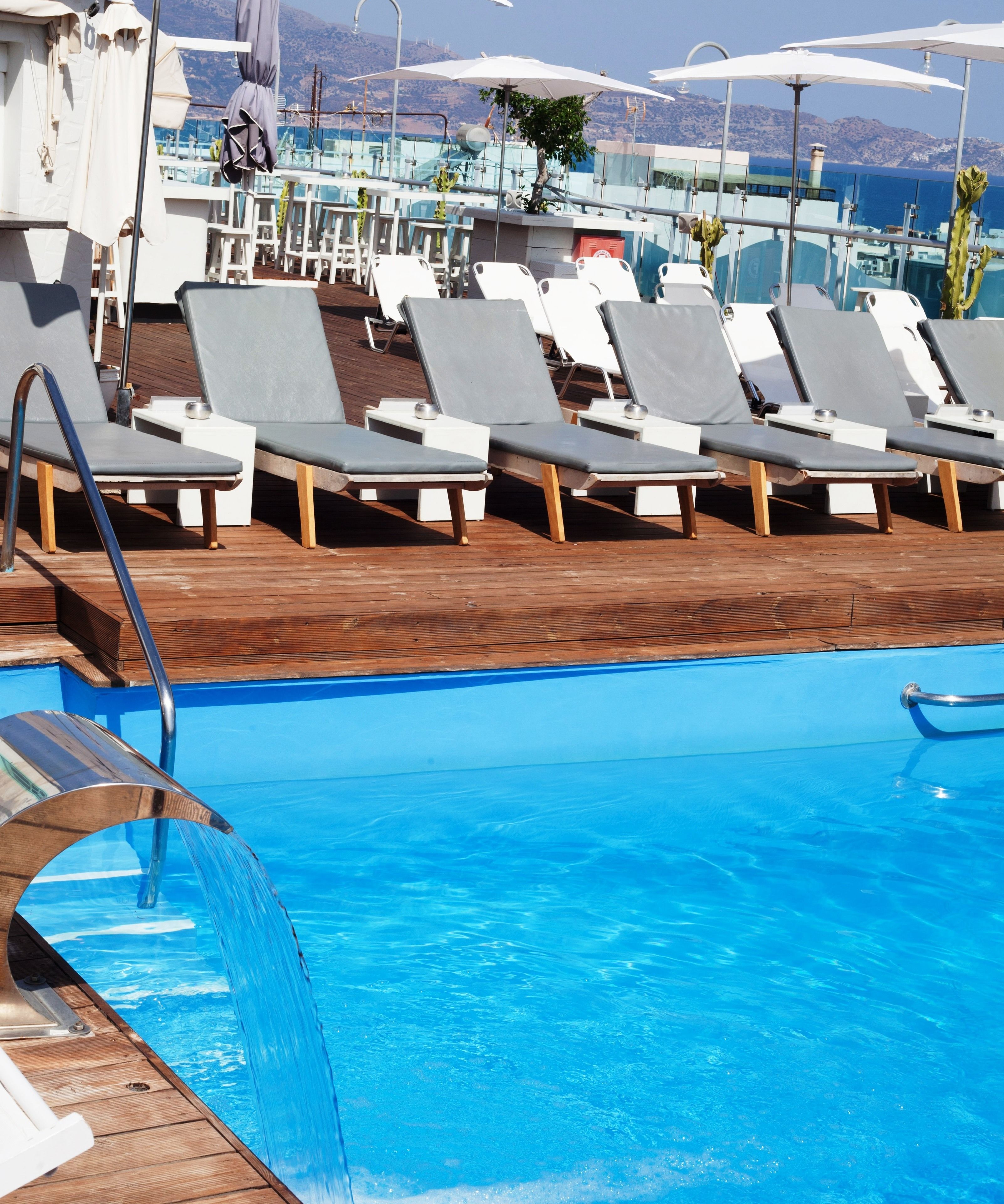 A poolside area with lounge chairs and umbrellas, a wooden deck, and a view of the sea.