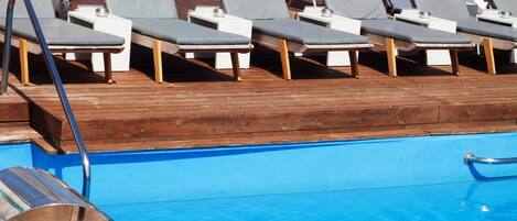 A poolside area with lounge chairs and umbrellas, a wooden deck, and a view of the sea.