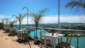 A rooftop terrace with tables and chairs, overlooking a cityscape and a body of water. - Capsis Astoria Heraklion Hotel (Heraklion)
