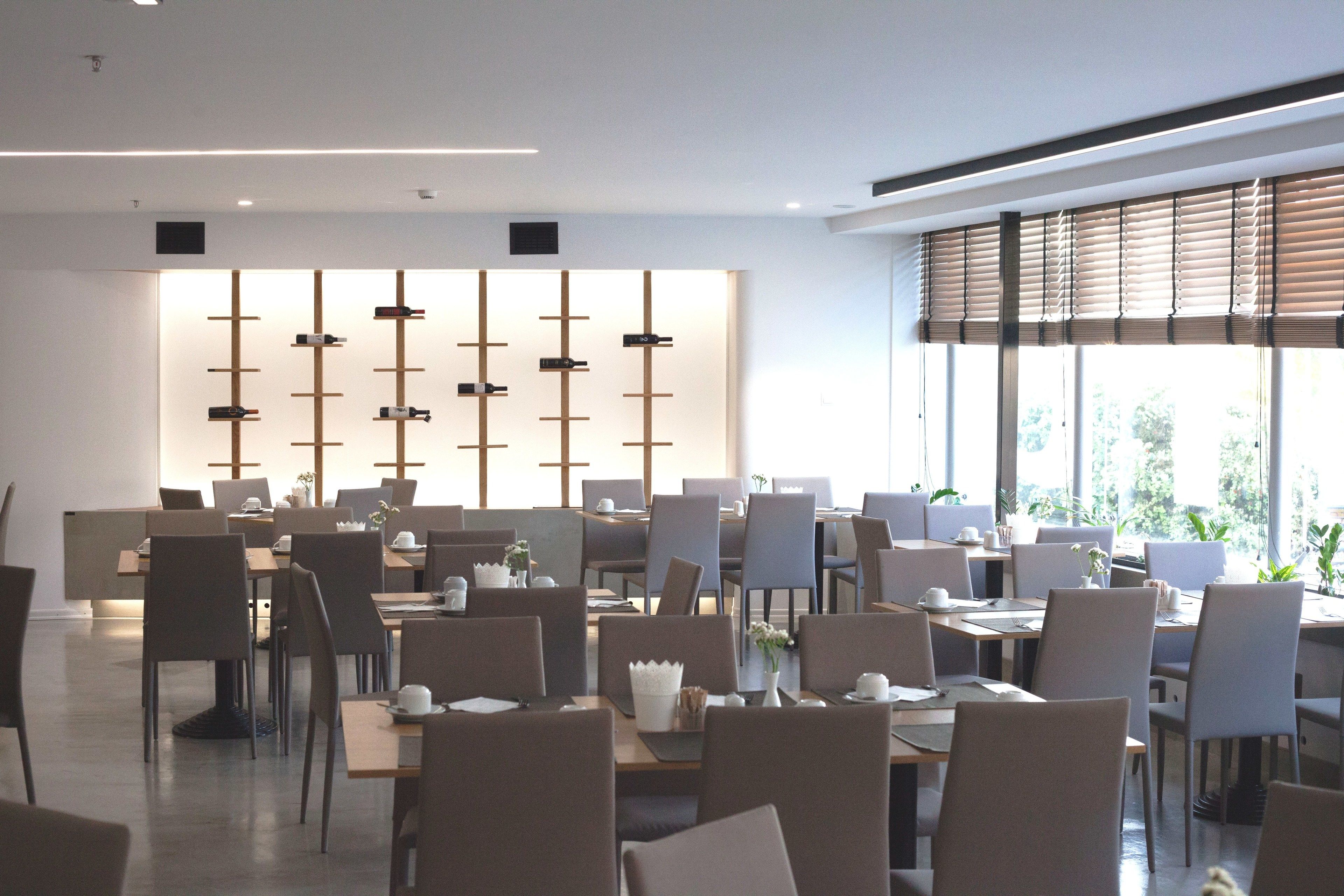 Un comedor moderno con varias mesas y sillas, una pared decorativa con elementos verticales y grandes ventanales con persianas.