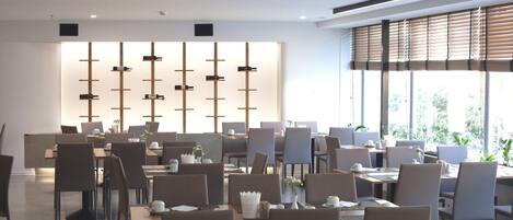 Un comedor moderno con varias mesas y sillas, una pared decorativa con elementos verticales y grandes ventanales con persianas.