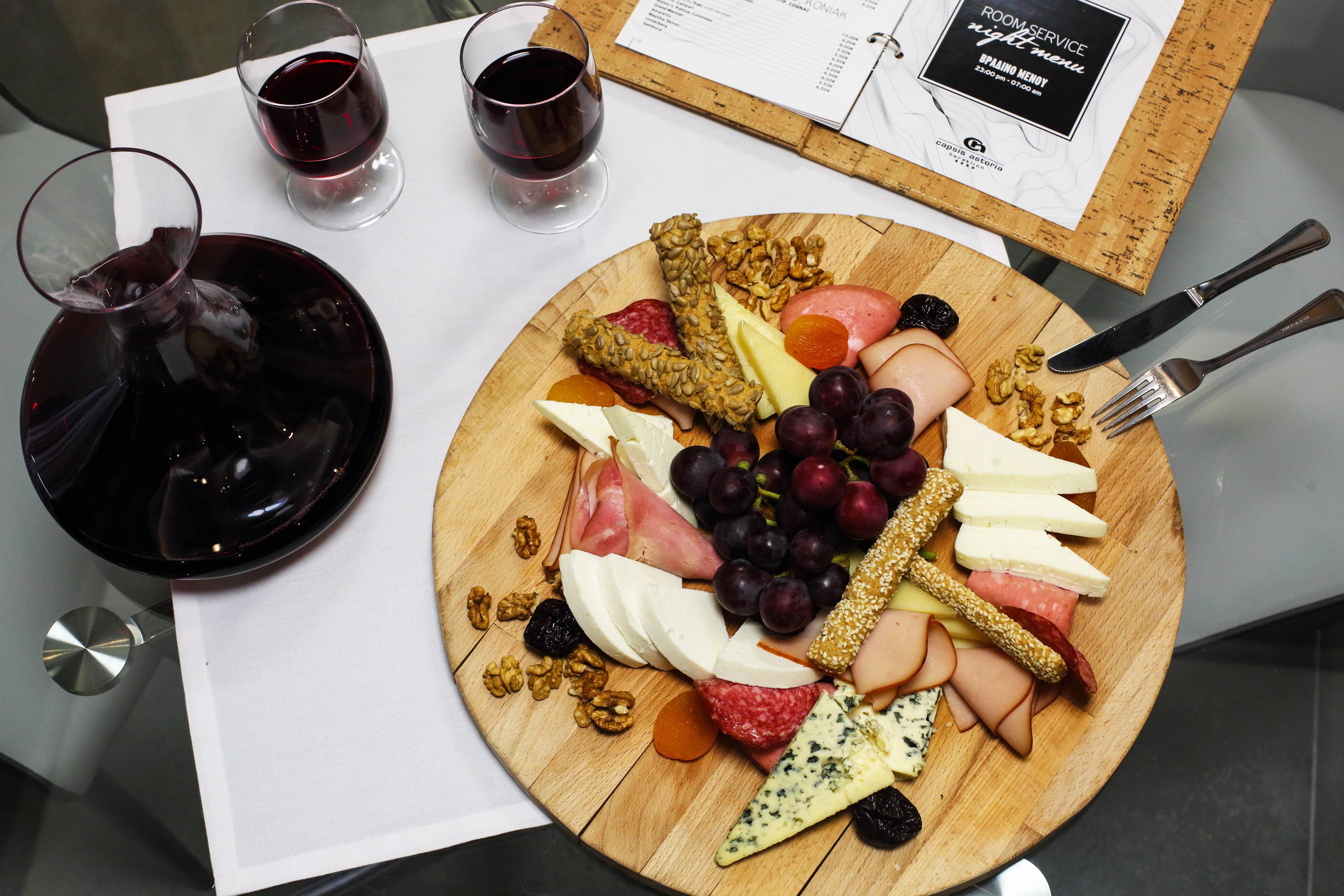 Una tabla de madera con varios quesos, frutas y frutos secos, acompañada de dos copas de vino tinto sobre una mesa con un menú y cubiertos.