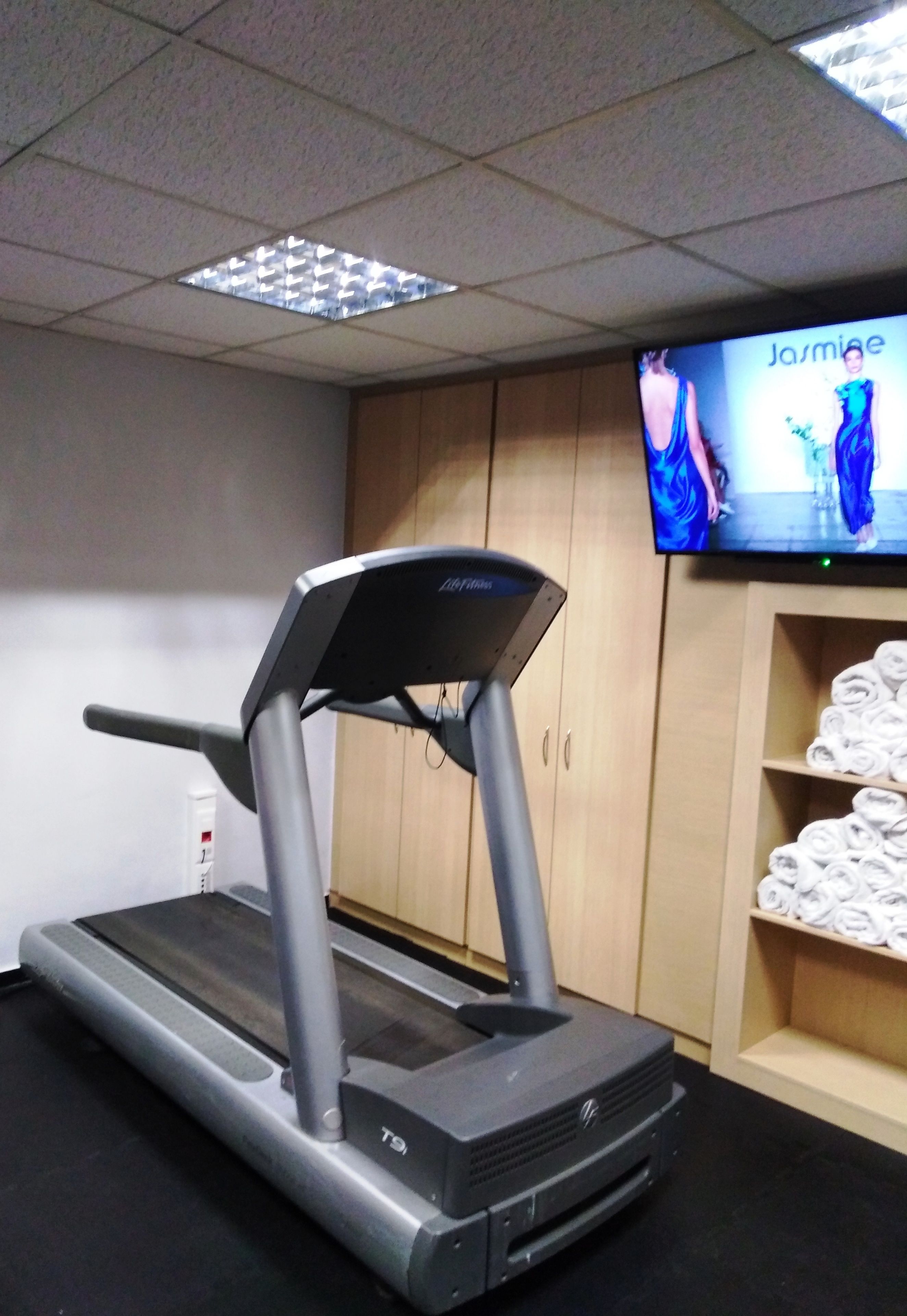 A treadmill in a room with a television displaying a fashion show, wooden cabinets, and a shelf with towels.